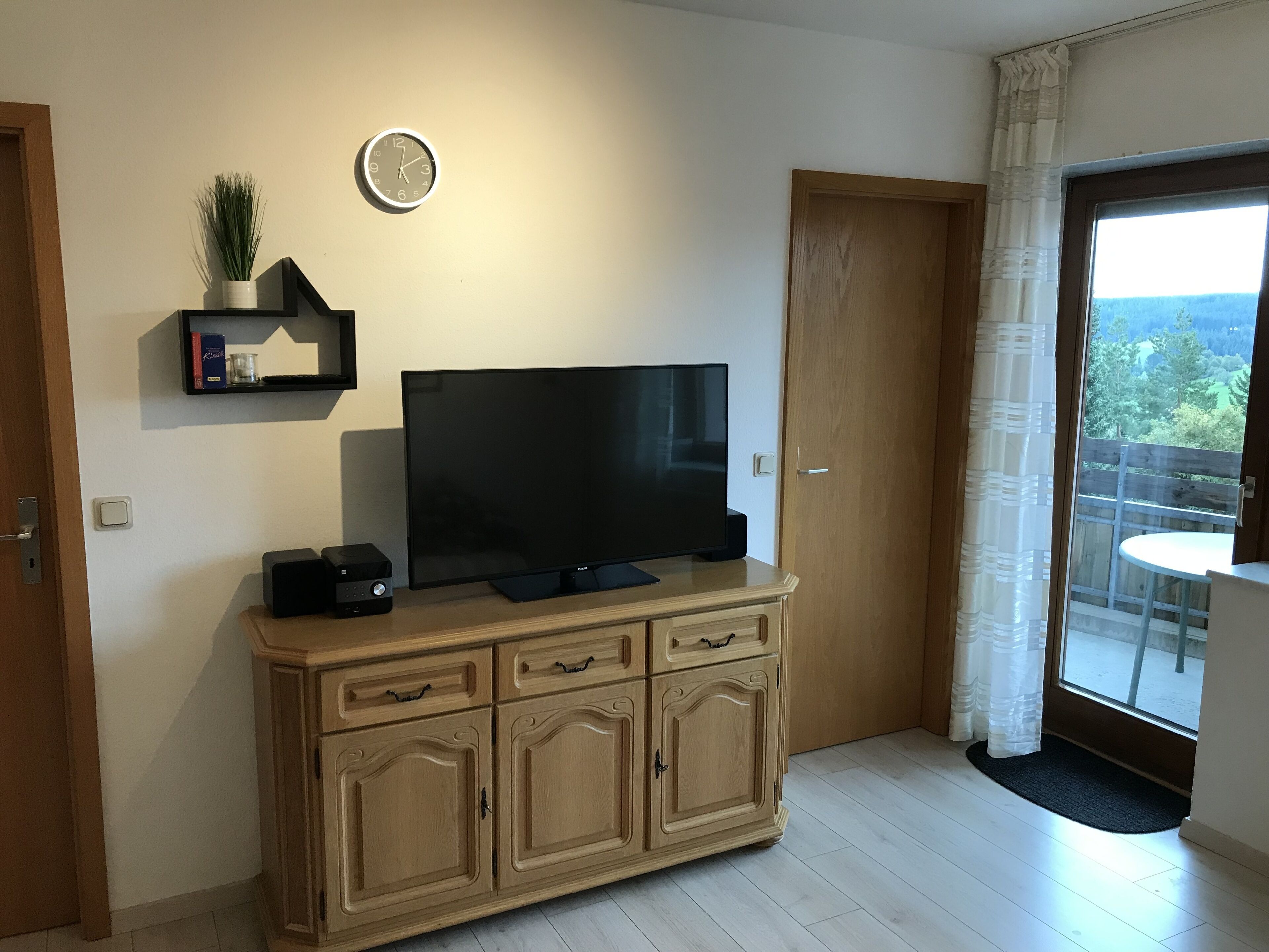 Un soggiorno con una TV a schermo piatto su un mobile in legno, un orologio sulla parete e una porta scorrevole in vetro che dà su un balcone.