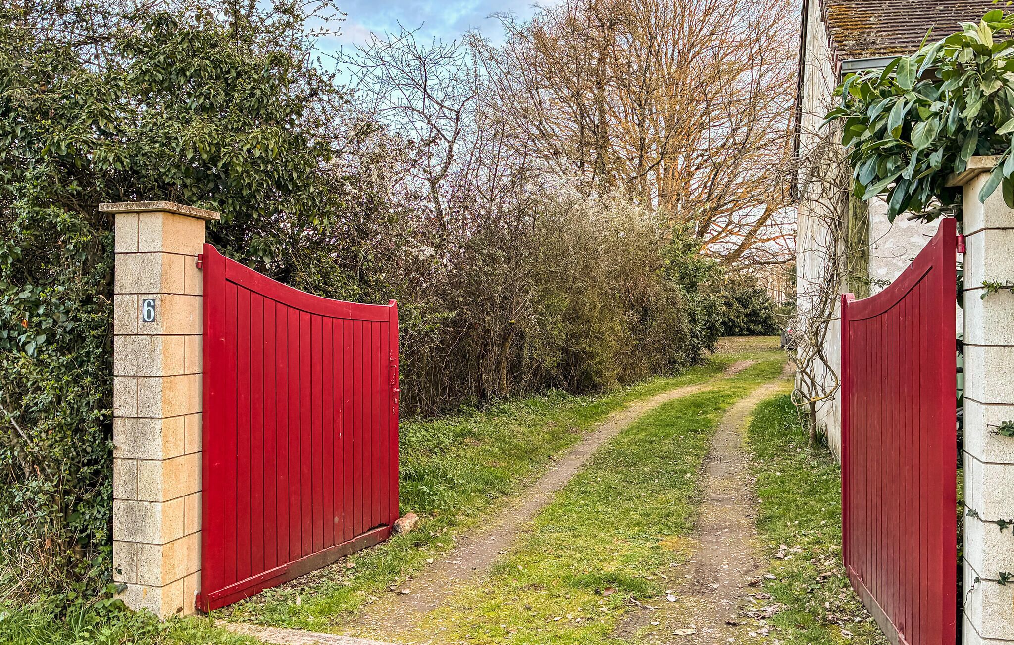Un étroit sentier mène à une porte rouge, entourée de verdure et bordée d’un bâtiment dont le mur est en pierre.
