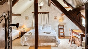 A cozy attic bedroom with a sloped ceiling, wooden beams, a bed with white linens, a nightstand with a lamp, and a small table with a chair. - Cozy home in Beauchene with WiFi (Beauchene)