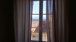 A window with curtains, revealing a view of a tiled roof and a distant landscape. - Casa Delia (L'Île-Rousse)