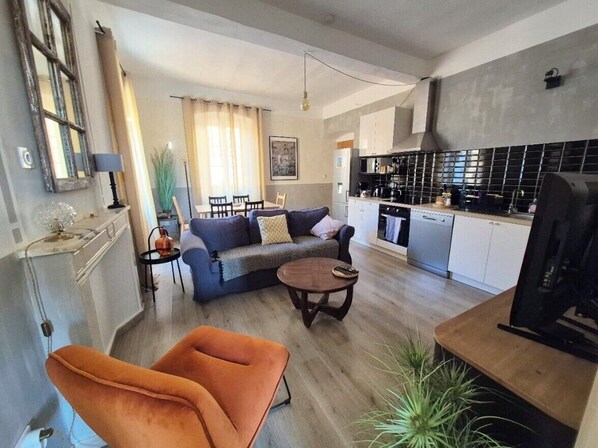 Un salon moderne avec un canapé, un fauteuil et une table basse, une cuisine avec un dosseret de carrelage noir, et un coin repas avec une table et des chaises.