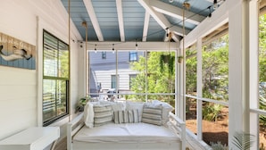A covered outdoor seating area with a white bench, a cushioned bench with pillows, and a view of a house and trees.
