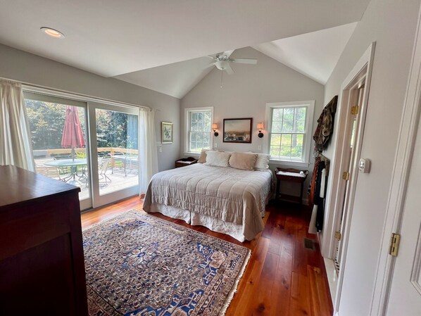 A bedroom with a large bed, a ceiling fan, a desk with a chair, and a view of the outdoors through a sliding glass door.