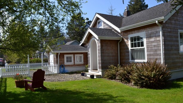 A two-story house with a front porch, a white picket fence, and a red Adirondack chair. - Adorable Cottage with Americana Theme, in the Heart of Gearhart. (Seaside)