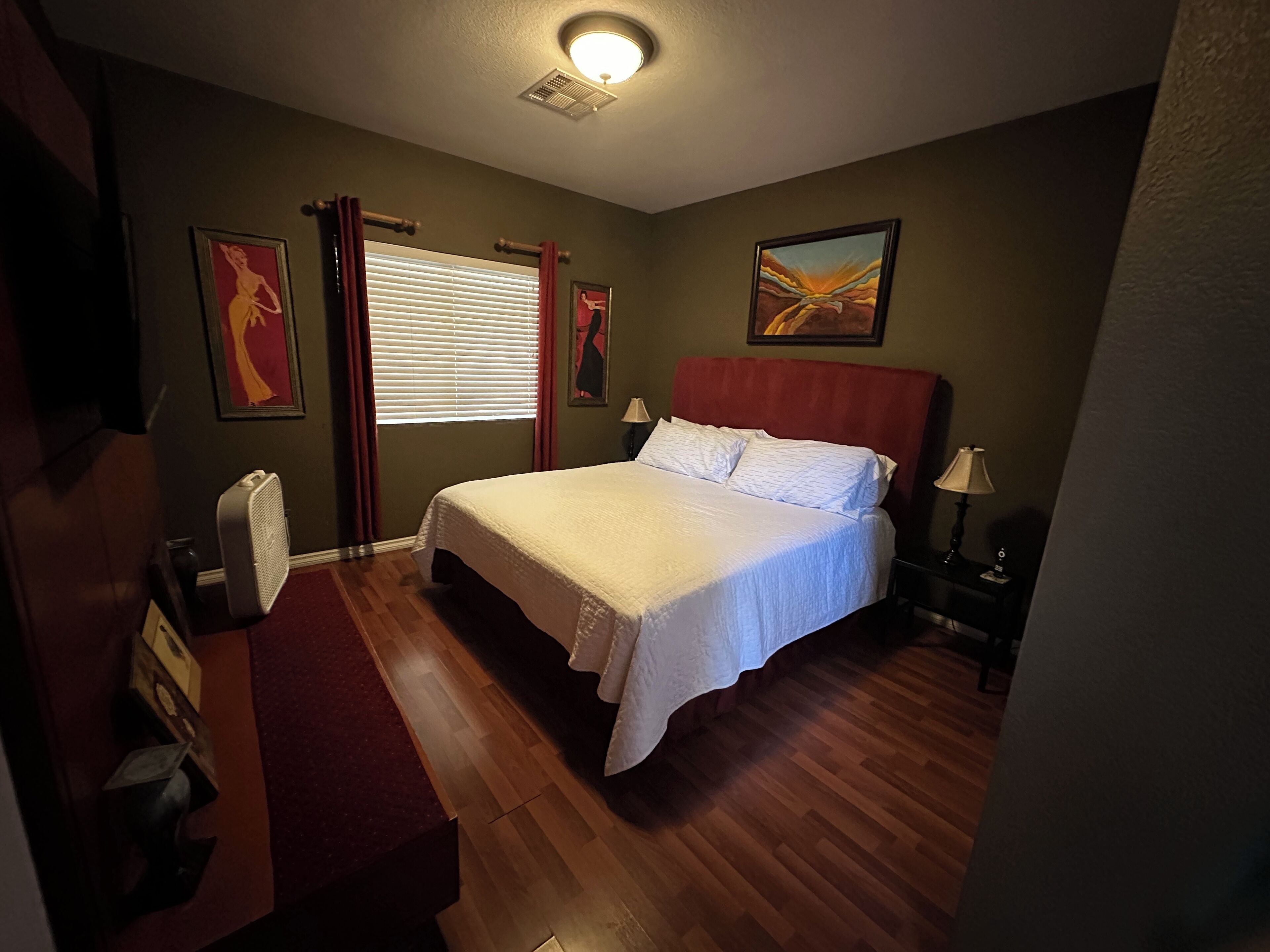 A bedroom with a bed, a window with curtains, a painting on the wall, and a ceiling light.