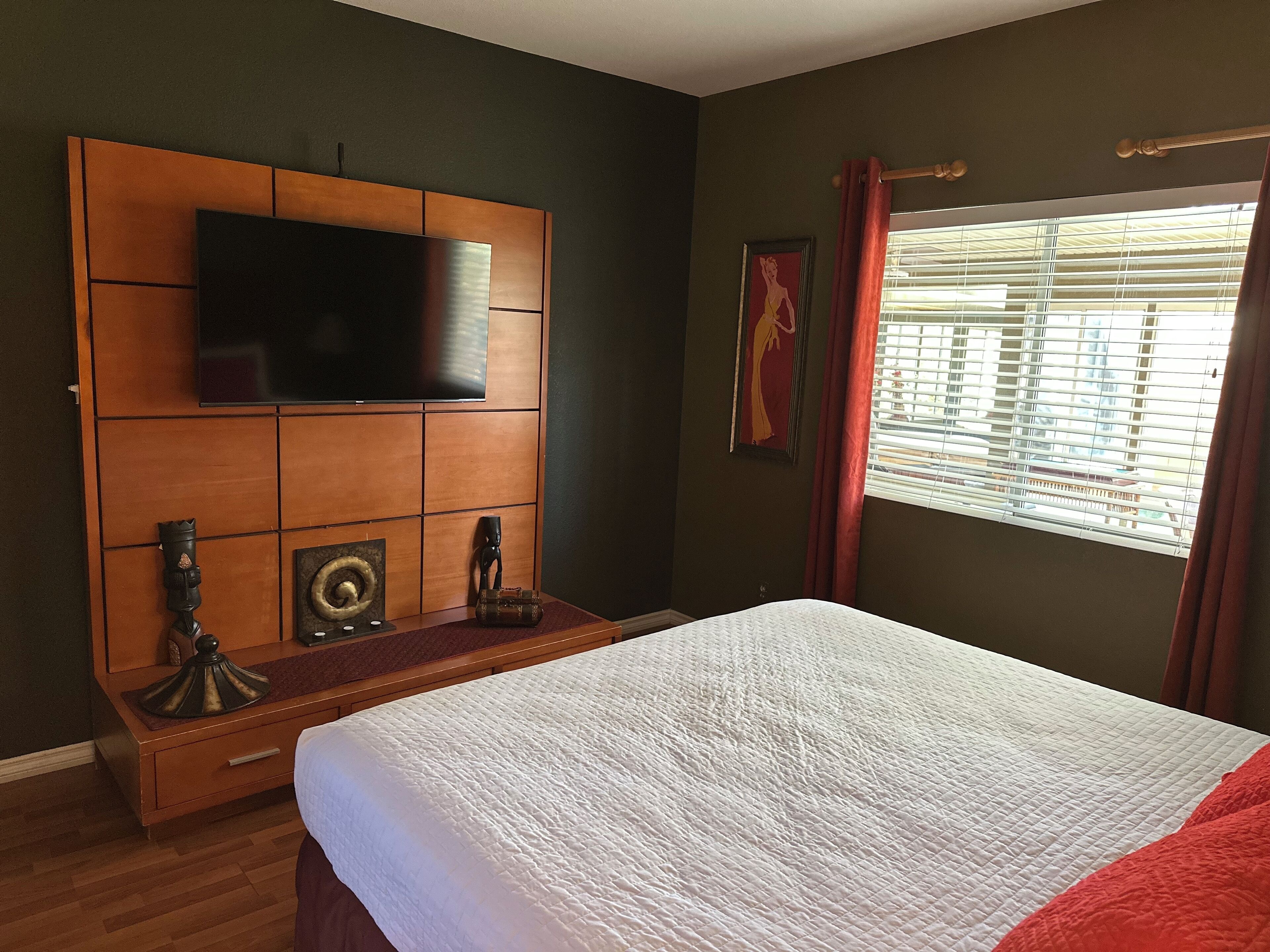 A hotel room with a large bed, a mounted TV, a wooden headboard, and a window with blinds.