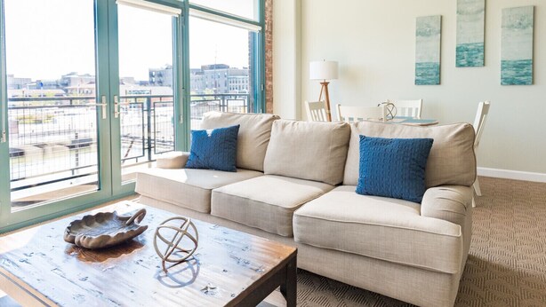 Ein Wohnzimmer mit einem beigefarbenen Ecksofa, einem Holztisch und Blick auf die Stadt durch große Fenster.