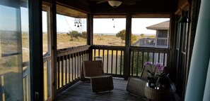 Eine Holzterrasse mit Korbstühlen und einem kleinen Tisch, mit Blick auf einen Strand und ein entferntes Gebäude.