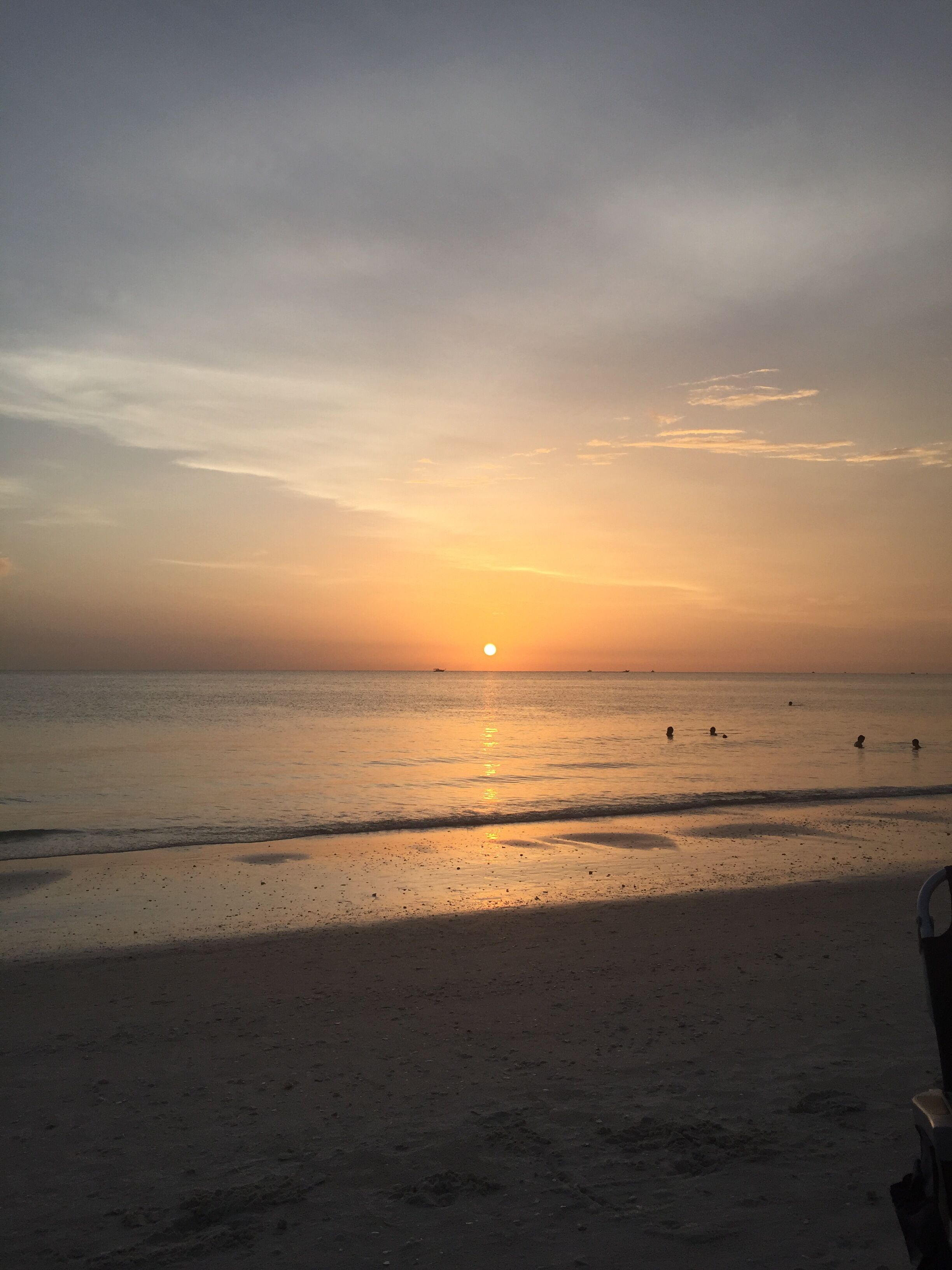 Une plage au coucher du soleil, avec une mer calme et quelques personnes qui se baignent.