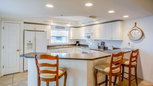 A modern kitchen with white cabinets, a central island with bar stools, a clock on the wall, and a window with a view of the ocean.