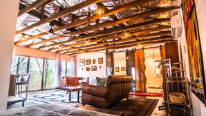 Una habitación amplia con techo alto, vigas de madera a la vista, un sofá de cuero, una mesa de comedor y un ventanal que proporciona luz natural.