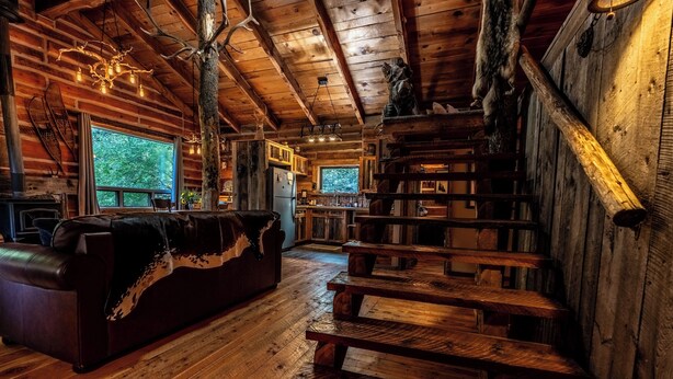 A rustic cabin interior with a wooden staircase, a leather sofa, and a large window overlooking greenery.