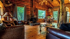 A rustic cabin interior with wooden walls, a stone fireplace, leather furniture, and large windows offering a view of the forest.