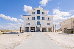 Un grand bâtiment blanc à plusieurs étages, doté d’une entrée centrale et entouré d’une zone sablonneuse plantée de palmiers.