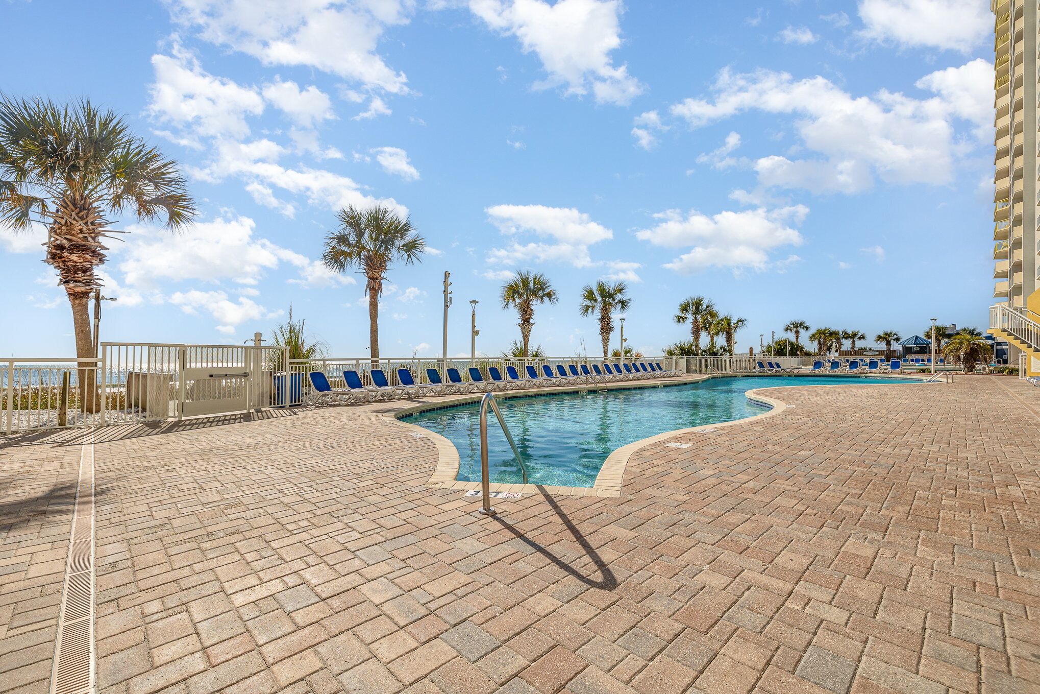 A pool area with a curved pool, blue lounge chairs, palm trees, and a clear sky.