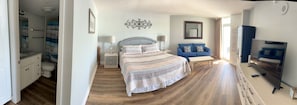 A bedroom with a bed, bedside tables, a nightstand, a blue sofa, a TV, and a bathroom with a shower curtain. - Oceanfront Breath Taking View with Private Balcony - 2bd/2ba Condo in NMB (North Myrtle Beach)