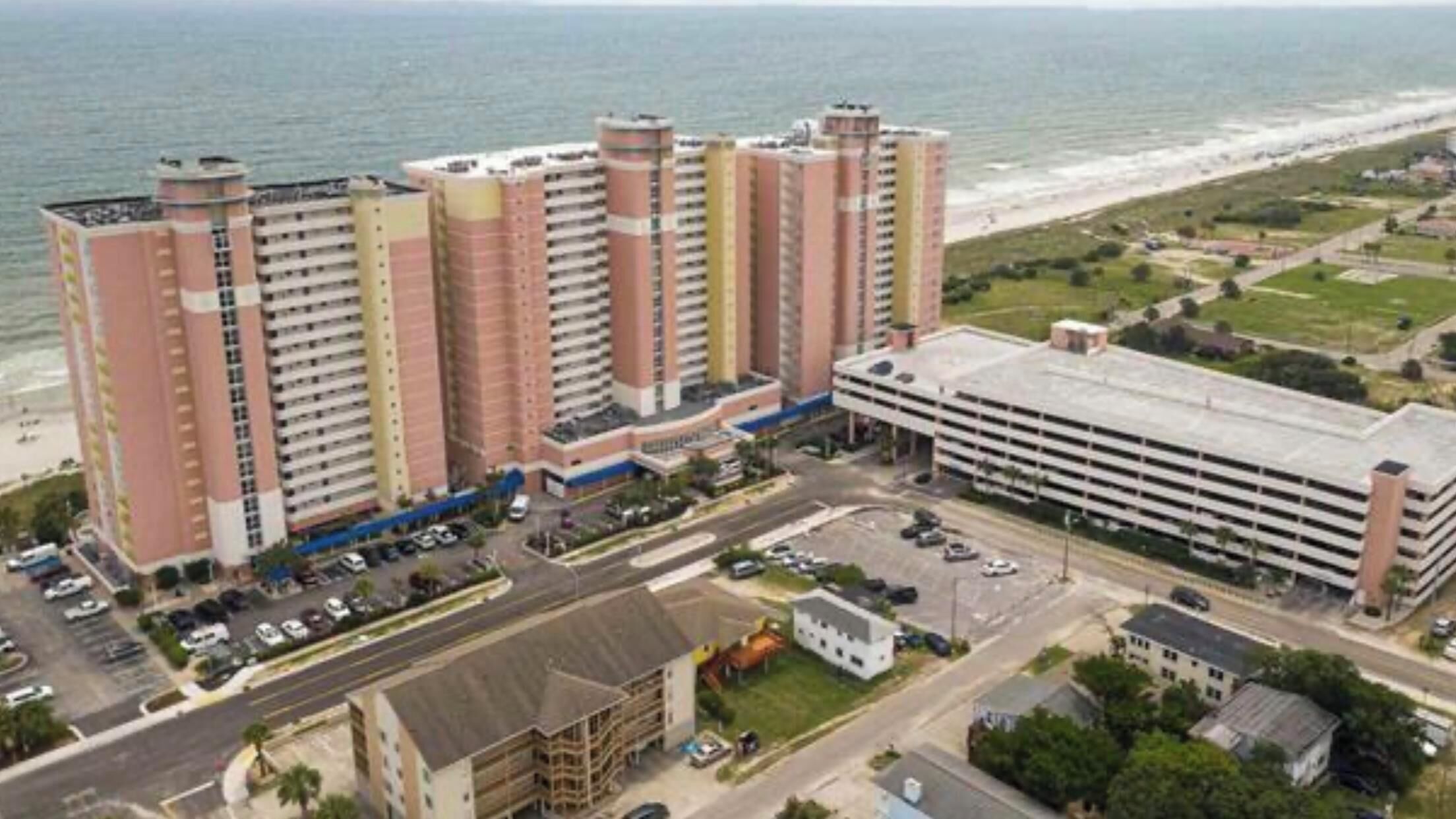 A high-rise hotel with multiple floors, a parking lot, and a view of the beach.