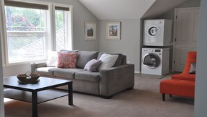 Un salon avec un canapé gris, un fauteuil rouge, un lave-linge et deux tableaux encadrés accrochés au mur.