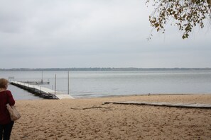A beach with a wooden pier extending into calm water, a sandy shore, and a distant tree line.