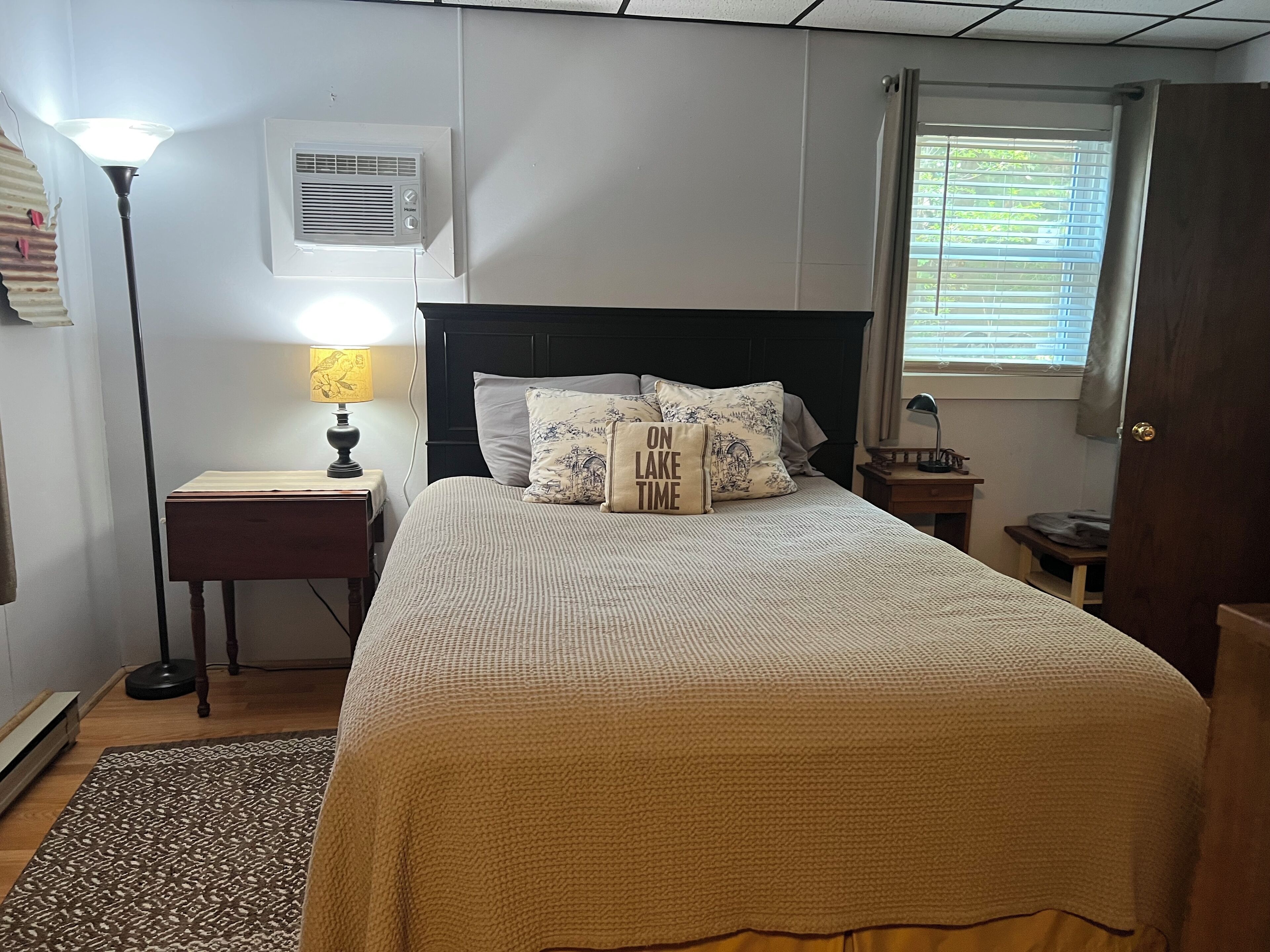 A neatly arranged bedroom with a bed, bedside lamps, a nightstand, a window with blinds, and an air conditioning unit on the wall.