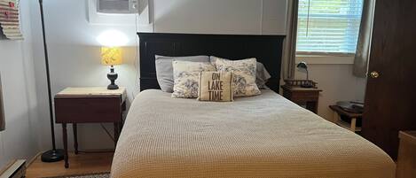 A neatly arranged bedroom with a bed, bedside lamps, a nightstand, a window with blinds, and an air conditioning unit on the wall.
