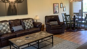 A living room with a brown leather sofa, a coffee table, a dining area with a chandelier, and a large moose artwork on the wall.