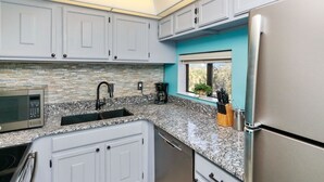 Une cuisine avec des meubles blancs, un plan de travail en granit, un réfrigérateur en acier inoxydable et une fenêtre donnant sur un paysage verdoyant.
