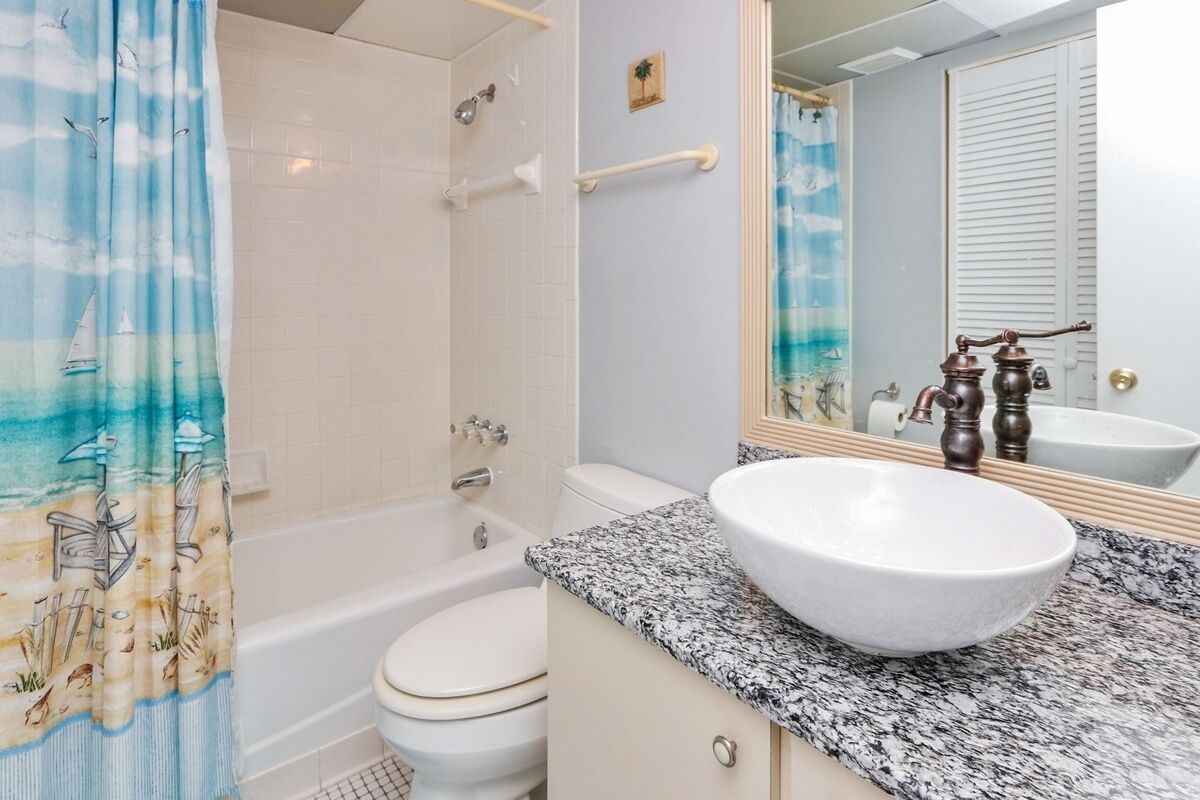 A bathroom with a white bathtub, a white toilet, a granite countertop with a white sink, and a shower curtain with a seagull and sailboat design.