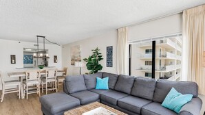 A living room with a grey sectional sofa, a coffee table, a dining area with a wooden table and chairs, a kitchen area, and a large window with curtains. - Gulf Front Corner Balcony - Sand Castle III 501 (Indian Shores)