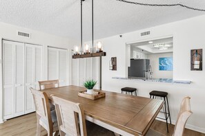 Un'area pranzo con un tavolo di legno, sedie, un lampadario e una cucina sullo sfondo, con soffitto bianco.