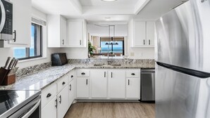 A modern kitchen with white cabinetry, a stainless steel refrigerator, and a view to the outside. - Gulf Front Corner Balcony - Sand Castle III 501 (Indian Shores)