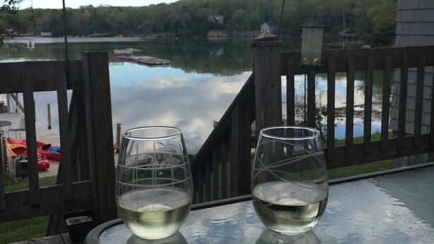 Two glasses of white wine on a glass table overlooking a lake with a wooden deck and a view of a house and trees in the background.