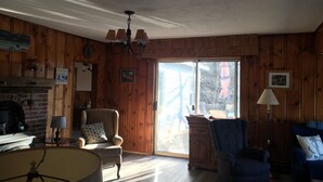 A cozy living room with wooden walls, a fireplace, a chandelier, and a large sliding glass door leading to a patio.