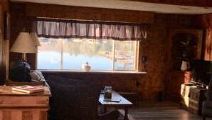 A cozy cabin interior with a large window overlooking a lake, a wooden desk with a lamp, and a chair.