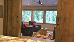 A bedroom with a large bed, two leather chairs with red cushions, a small wooden table, and a view of trees through large windows.