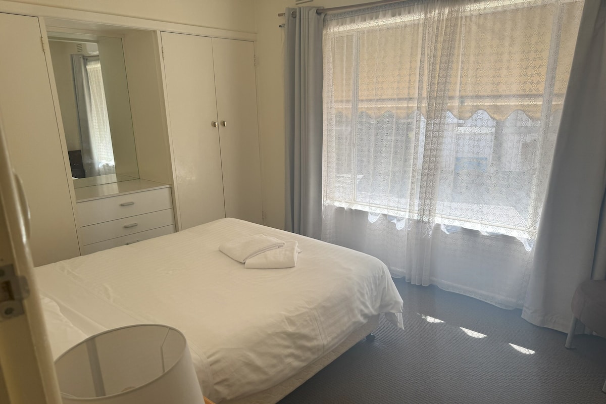 A hotel room with a neatly made bed, a bedside lamp, a wardrobe, and a window with sheer curtains.