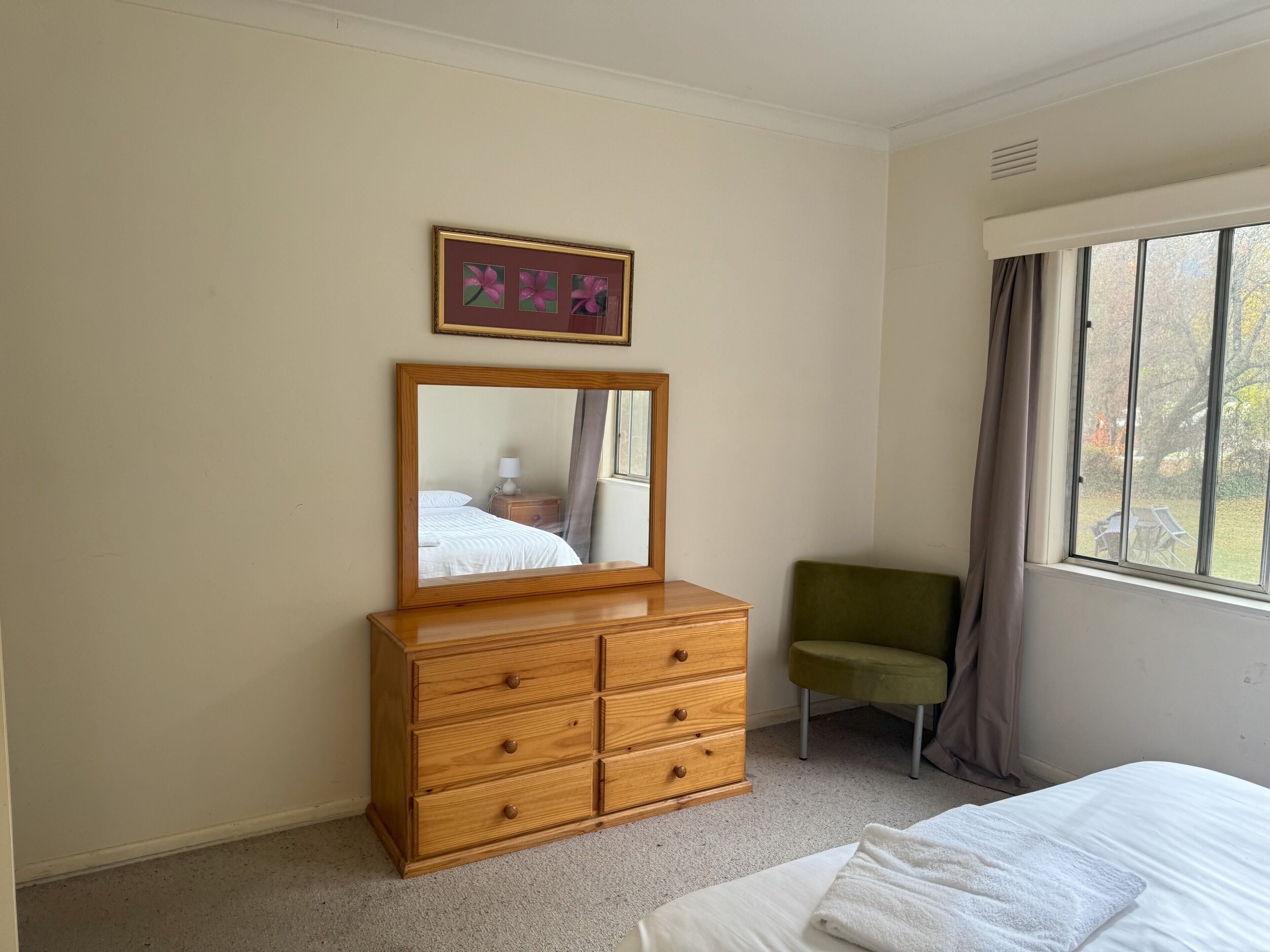 Un dormitorio con un aparador de madera, una silla verde, una cama, un ventanal con cortinas y un cuadro enmarcado en la pared.