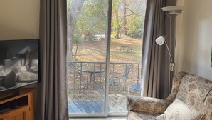 Un salon avec un téléviseur posé sur un meuble en bois, un canapé à motifs, une lampe de table et une porte-fenêtre coulissante donnant sur un espace extérieur avec une table et des chaises de patio.