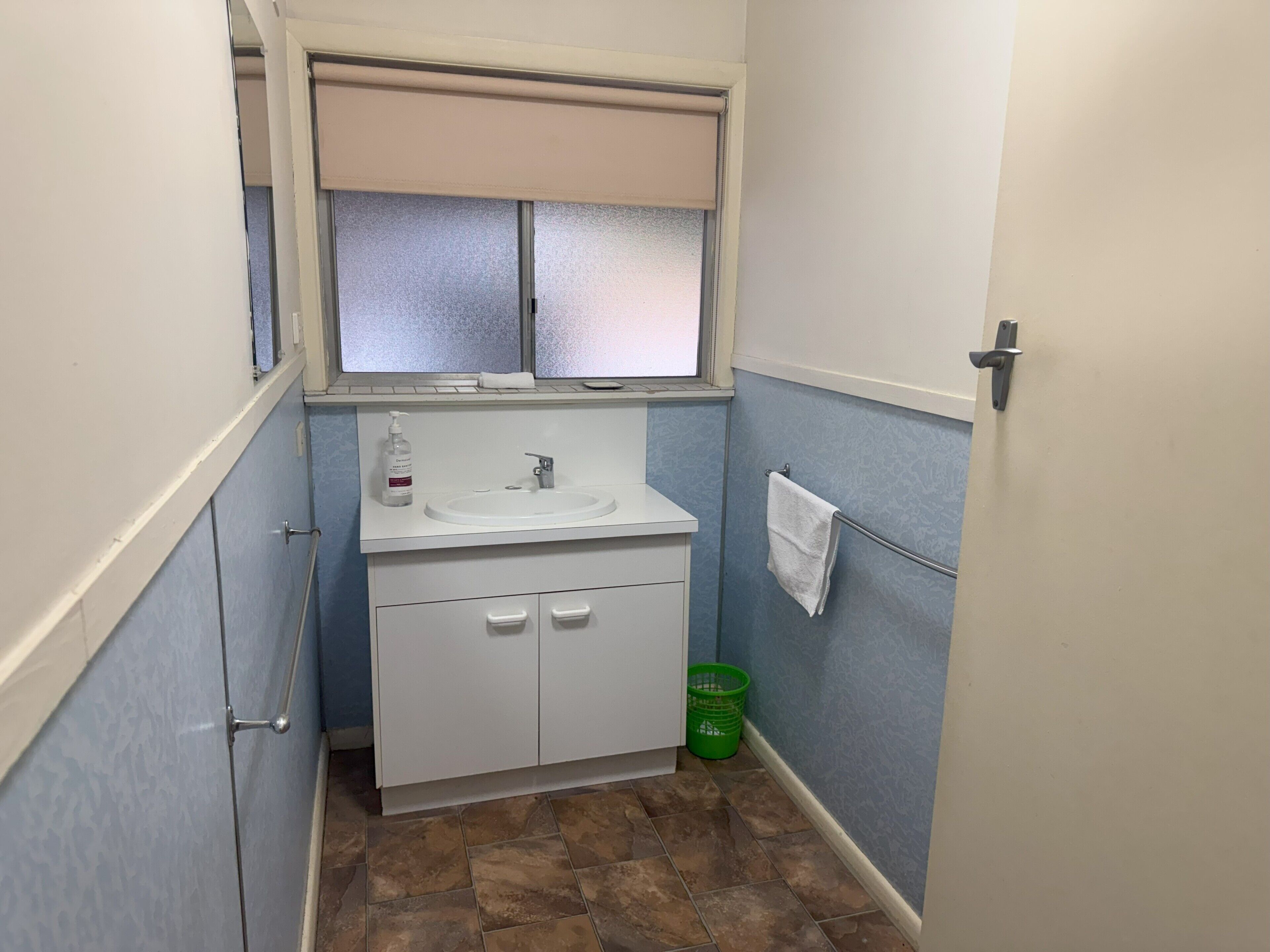 Ein kleines Badezimmer mit Waschbecken, einem Fenster mit Jalousien, einem grünen Korb und einem Handtuch an einem Halter.