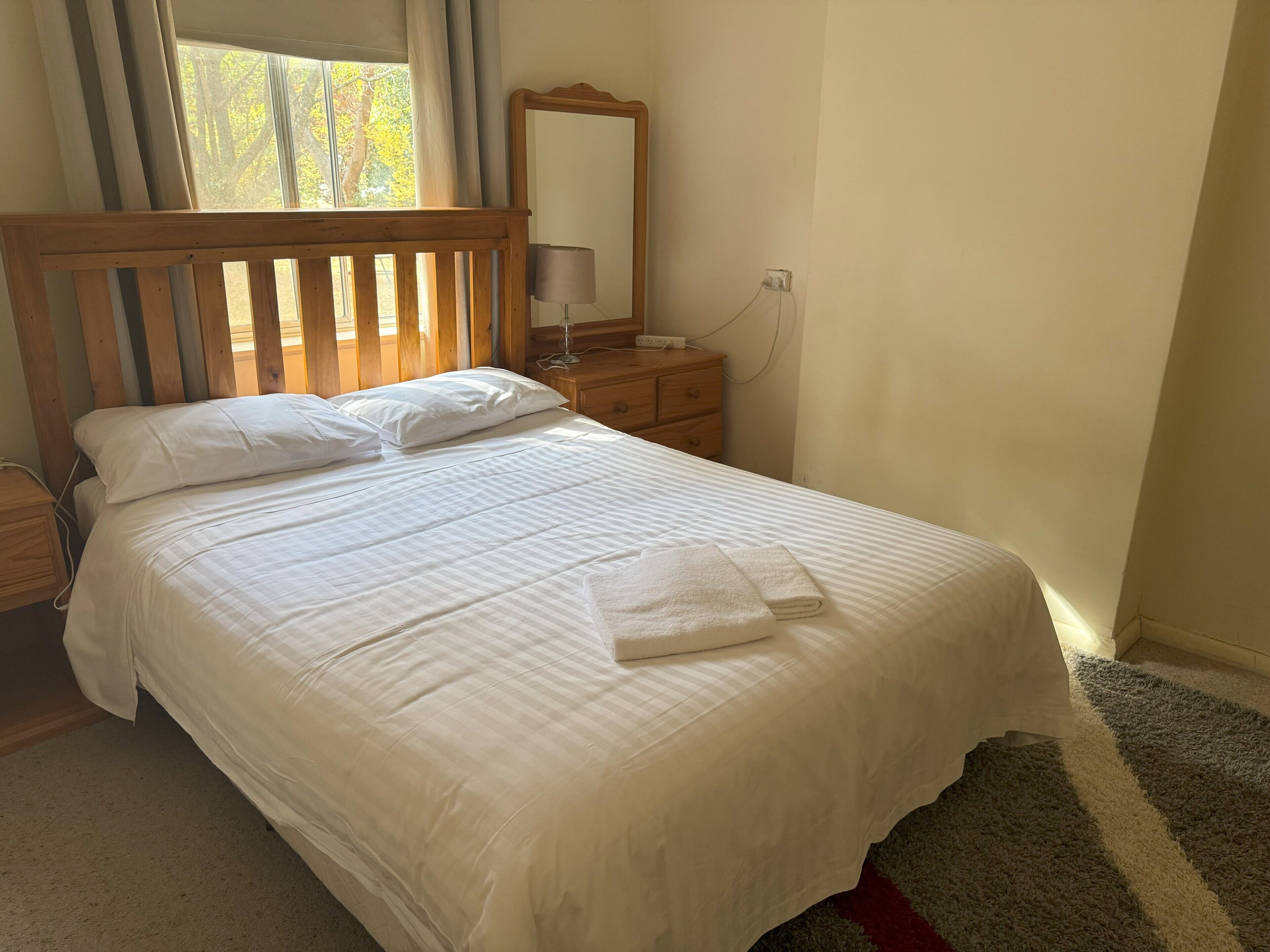 Een netjes opgemaakt bed met witte lakens, een houten hoofdbord, een nachtkastje met een lamp en een spiegel aan de muur.