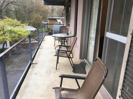 Un balcón con mesa y sillas, una silla de mimbre, y una vista a árboles y un edificio al fondo.