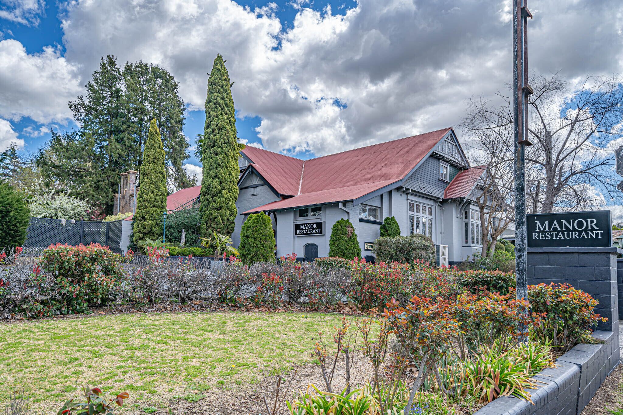 A manor restaurant with a red roof, surrounded by well-maintained gardens and tall trees.