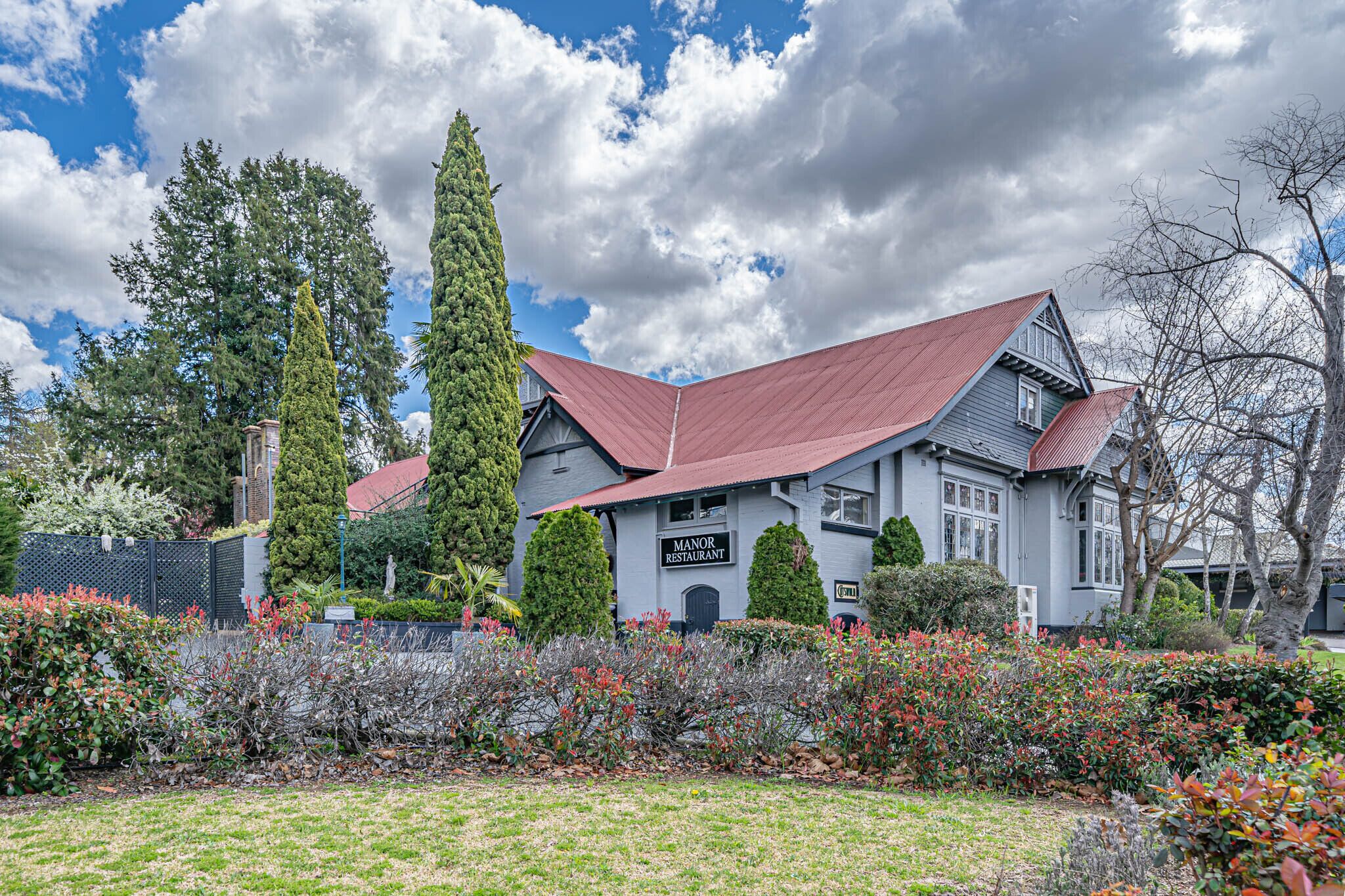 A two-story building with a red roof, labeled "Manor Restaurant," surrounded by well-maintained gardens and tall trees.