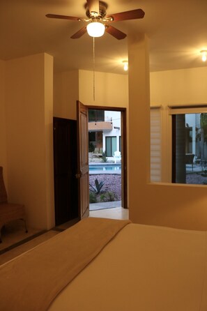 Una habitación con ventilador de techo, una puerta que da a un balcón y vistas a una piscina y un edificio.