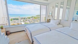 A bedroom with two beds, a large window with a view of a coastal town, and a small plant on the windowsill. - Coral garden 7 pools (Yomitan)