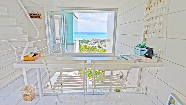 A compact room with a window overlooking a beach, a small table with a chair, and a wall-mounted shelf with decorative items. - Coral garden 7 pools (Yomitan)