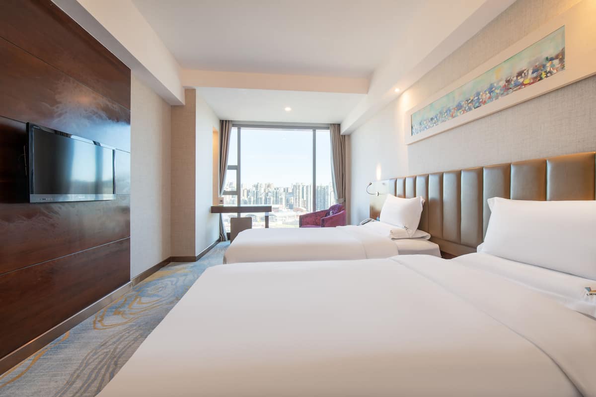 A hotel room with two beds, a large window with a city view, a flat-screen TV, and a painting on the wall.