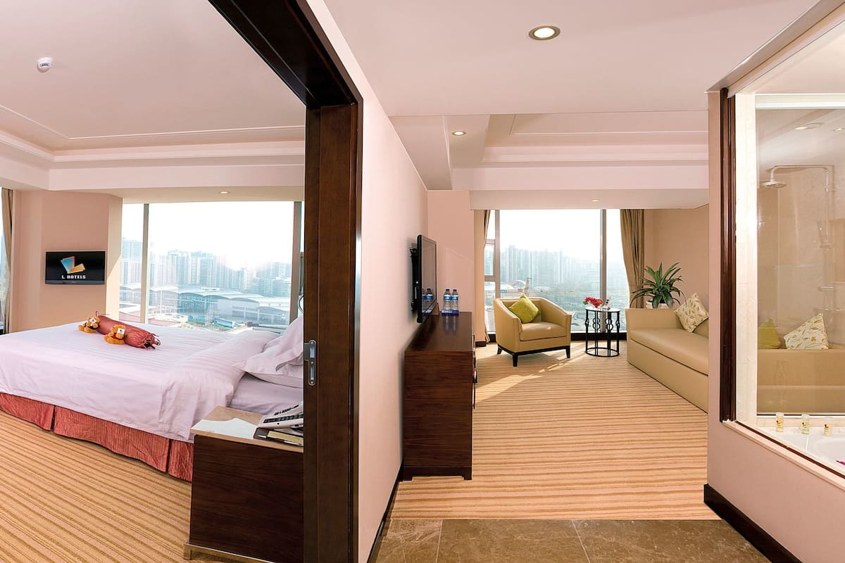 A hotel room with a large bed, a desk, a chair, and a sofa. There is a large window with a view of the city.