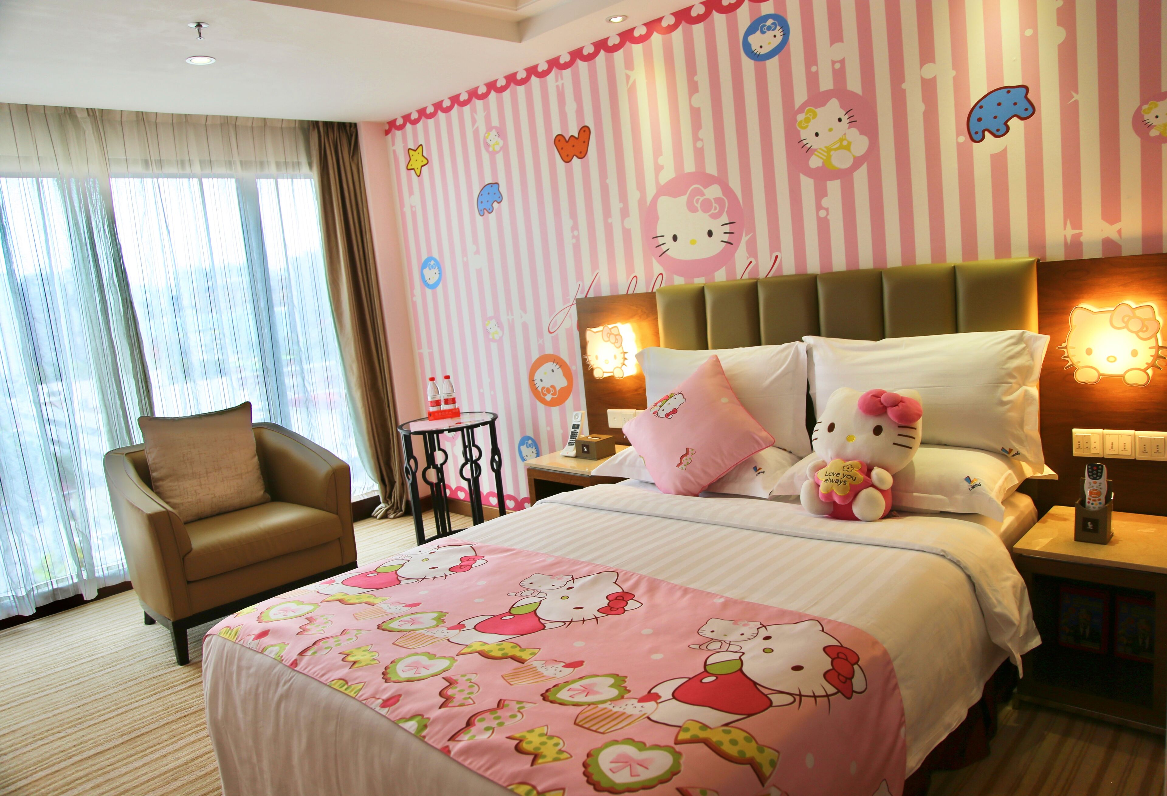 A hotel room with a bed featuring Hello Kitty decorations, a chair, a bedside table with a lamp, and a window with curtains.