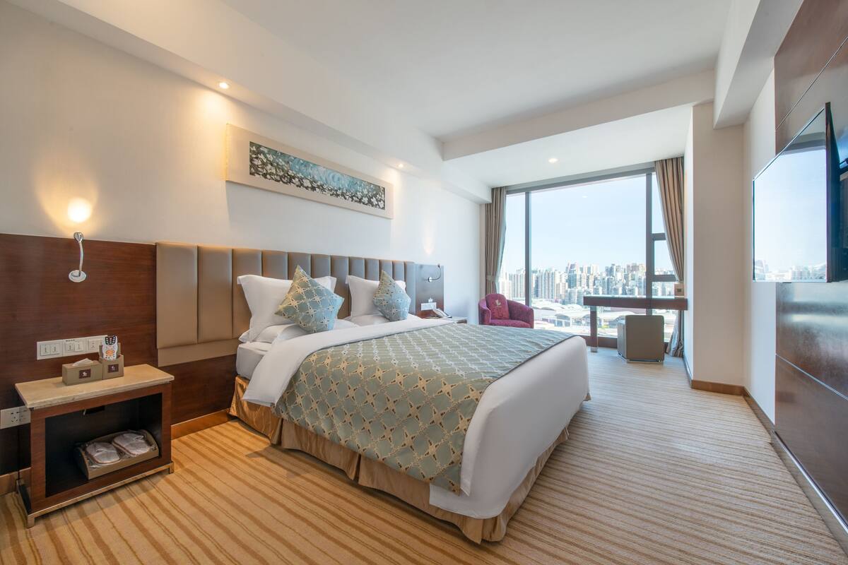 A modern hotel room with a large bed, a nightstand, a TV, and a city view.